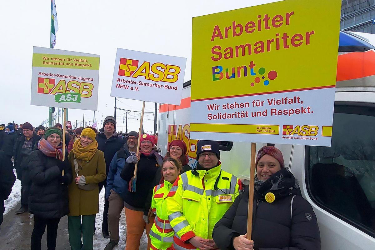 Die ASB-Mitarbeitenden und ihre Plakate vor einem Sanitätdienstfahrzeug des ASB Köln bei der Demonstration. 