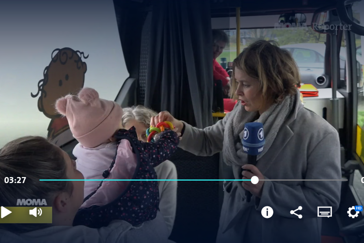 Die Reporterin gibt einem Baby im Hebammenmobil einSpielzeug. Im Hintergrund die Hebamme und die Rettungssanitäterin. 